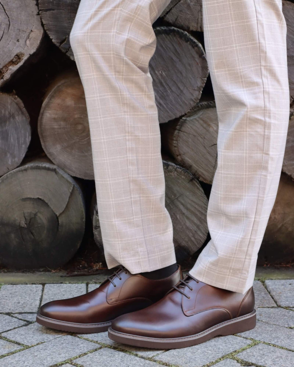Person wearing brown leather shoes and beige checkered pants standing in front of stacked logs.