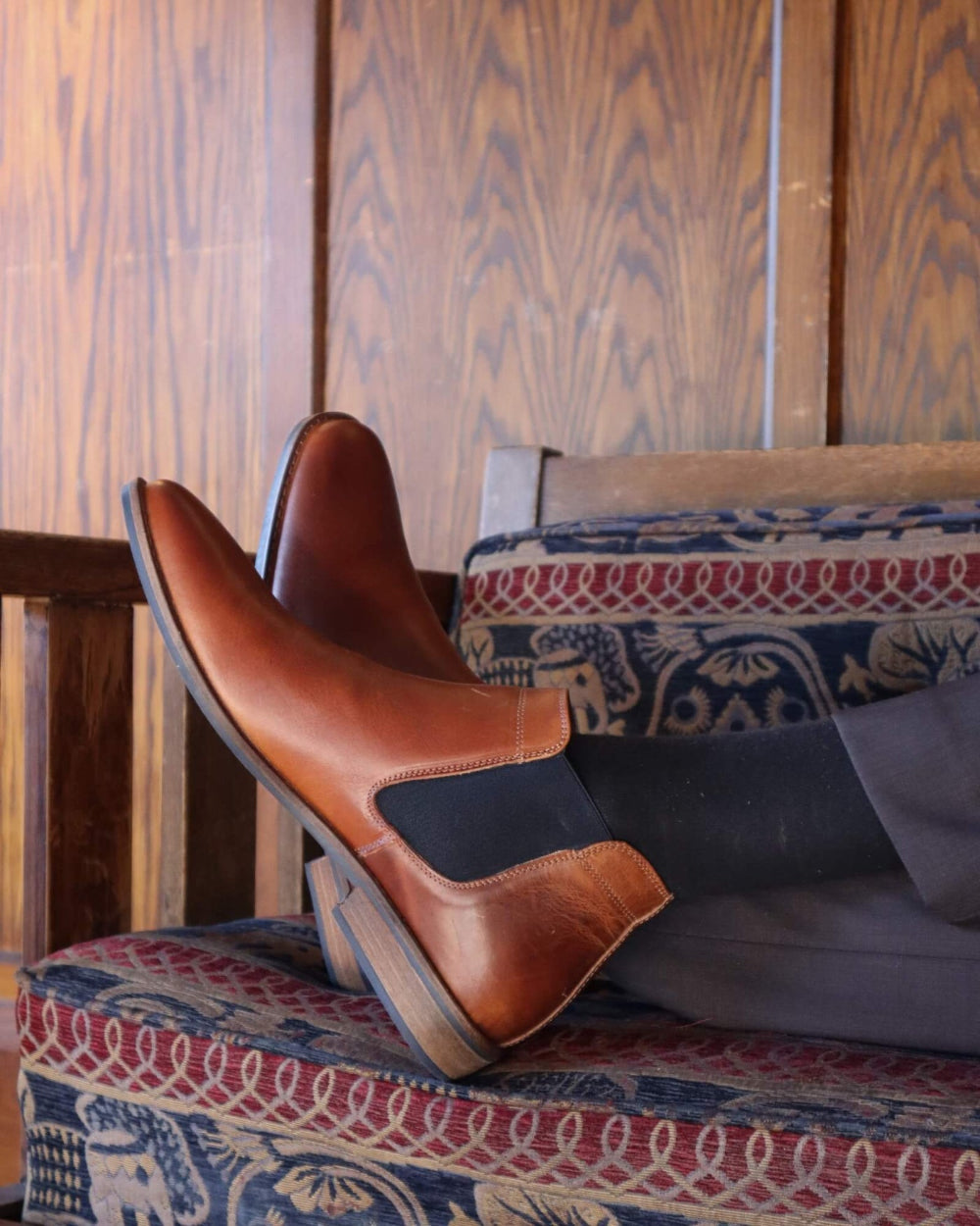 Brown leather shoe with a black patch on a patterned cushion against a wooden wall.